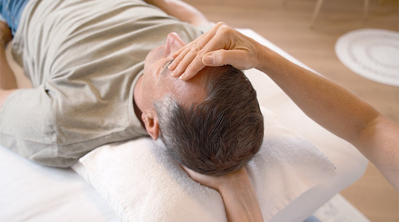 Reiki-Behandlerin hält die linke Hand unter den Kopf eines Kunden, der vor ihr auf der Liege liegt, die rechte Hand liegt auf der Stirn des Kunden