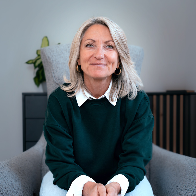 LEBENSKOMPONISTIN® Barbara Schwab-Melcher sitzt in ihrem grossen grauen Sessel mit Händen auf ihrem Schoß, leicht aufmerksam nach vorne gebeugt, mit lächelndem klarem Blick direkt in die Kamera